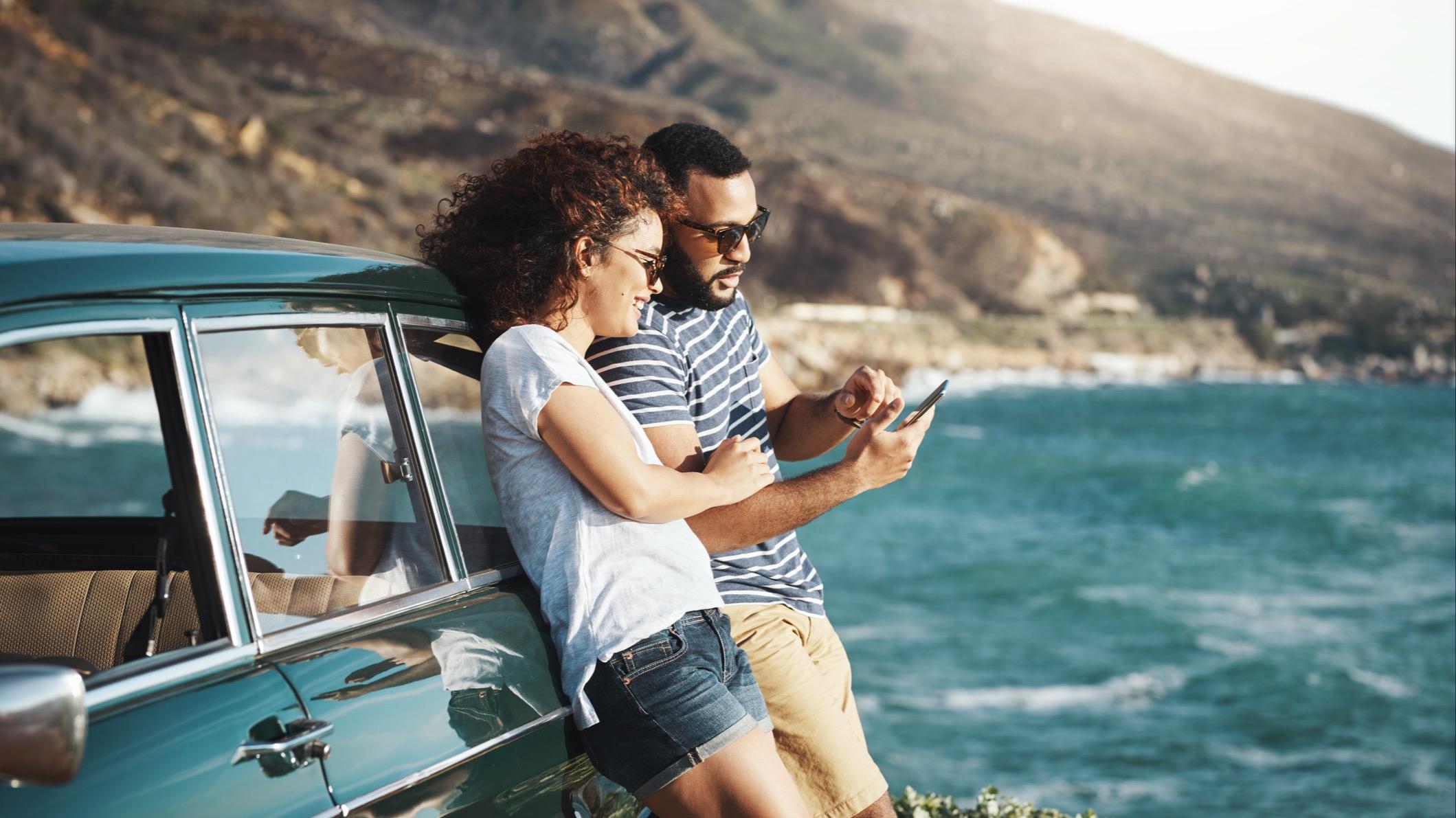 Man and woman looking at a smartphone while leaning against a car by a body of water