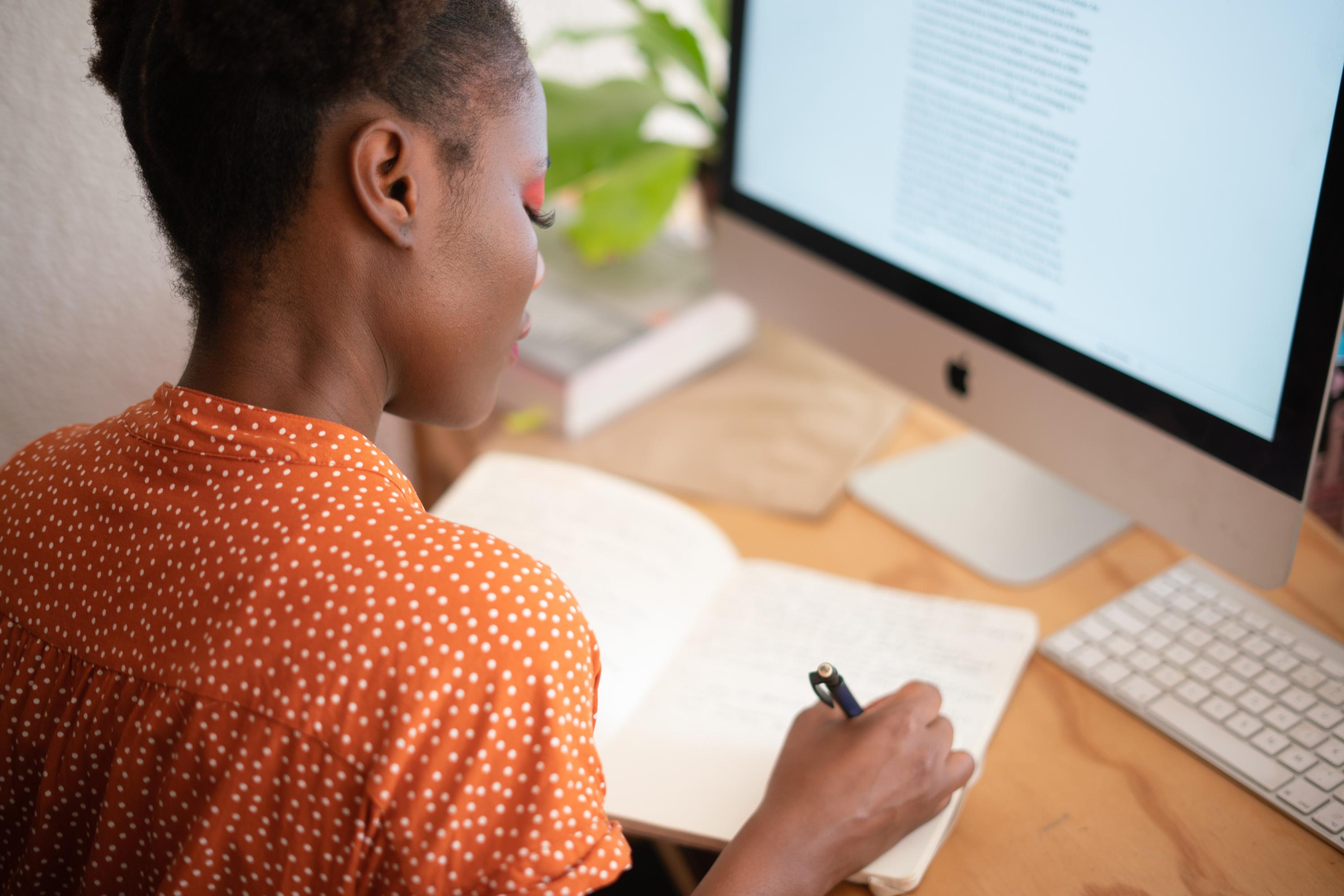 Woman writing in a notebook in front of a computer monitor